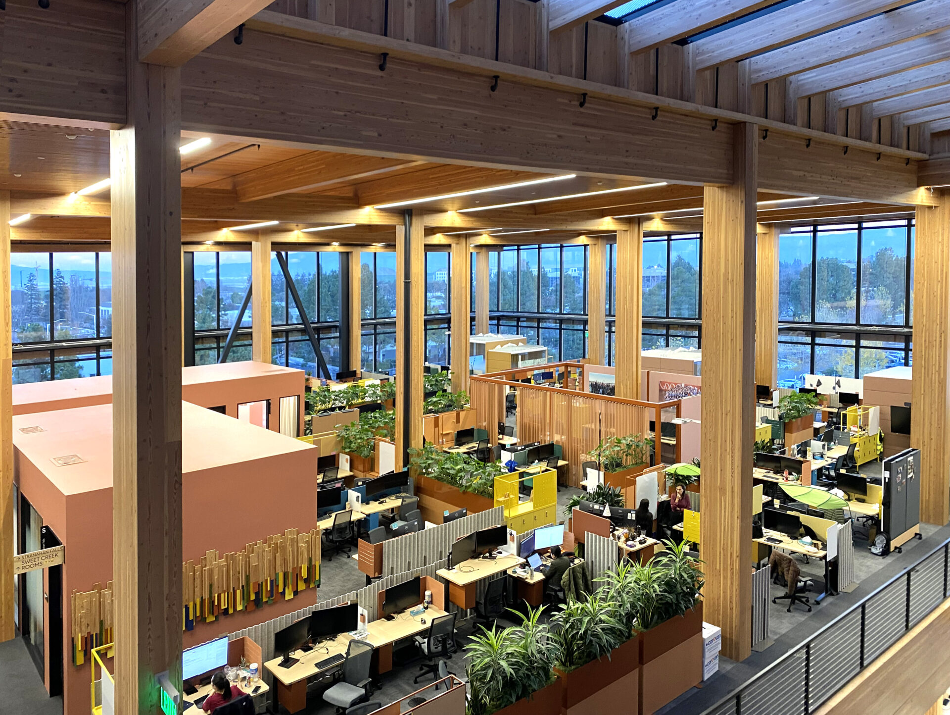 An overhead view from an upper floor of an office space with cubicles, desks, and computers with floor to ceiling windows and beams of timber supporting the building