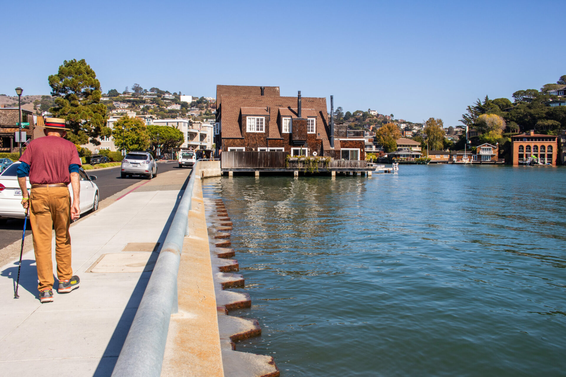 Elderly man with cane and hat walking with back to viewer down a sidewalk beside a calm body of water nearly level with the sidewalk.