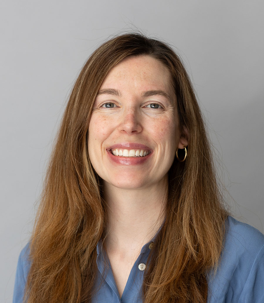 Headshot of a white woman with reddish-brown hair, gold hoop earrings and a blue button down shirt