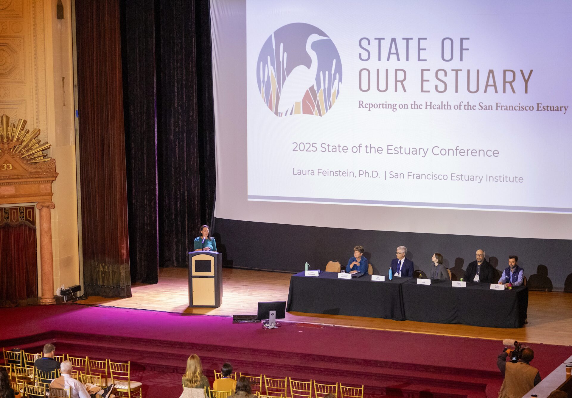 A panel sits at a table on an auditorium stage with a presentation in the background and a slide saying "State of Our Estuary: Reporting on the Health of the San Francisco Estuary"