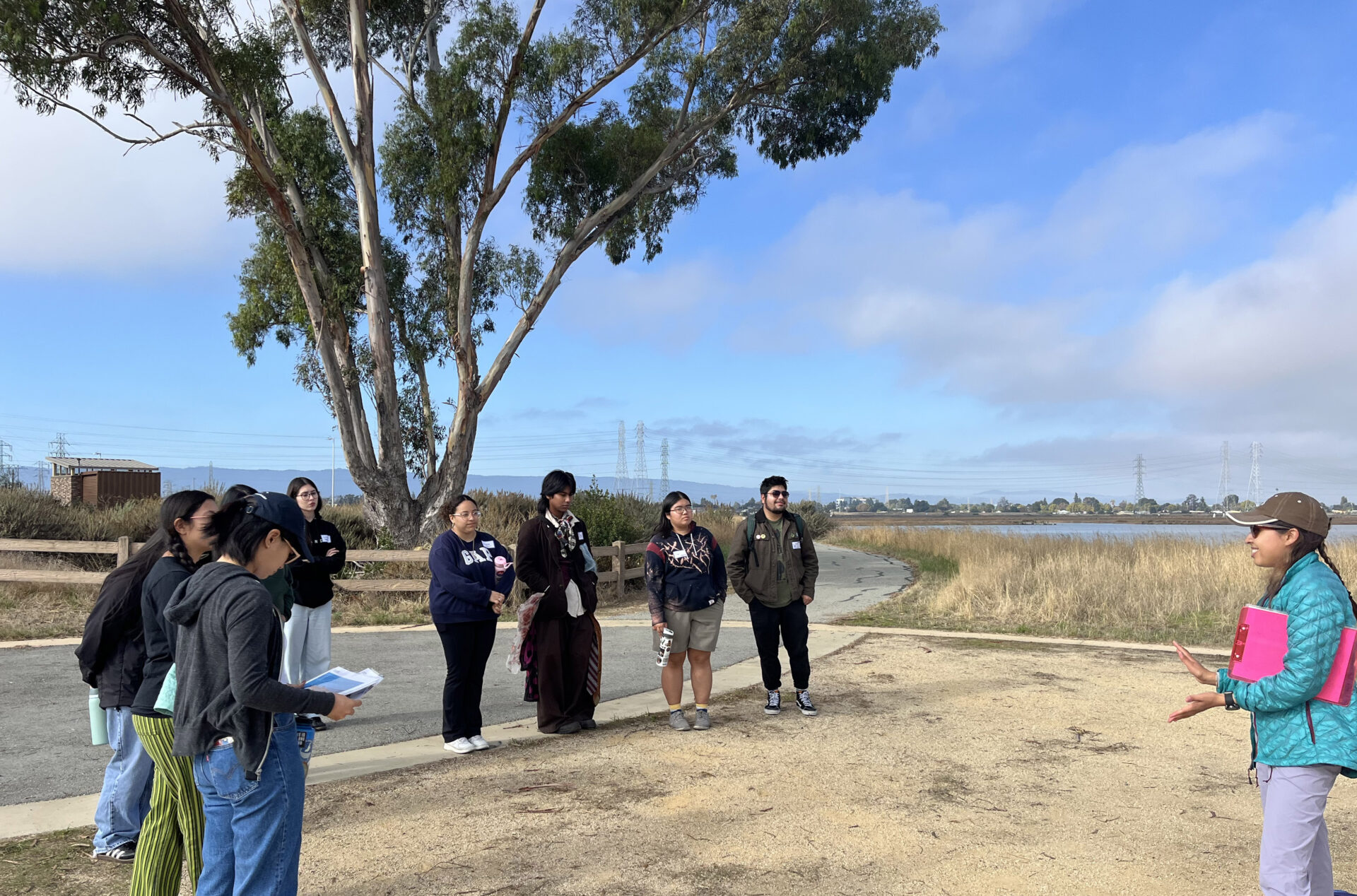 A woman in a blue jacket, a brown hat, purple pants and carrying a clipboard speaks to a group of about eight students in front of a large tree and a marsh