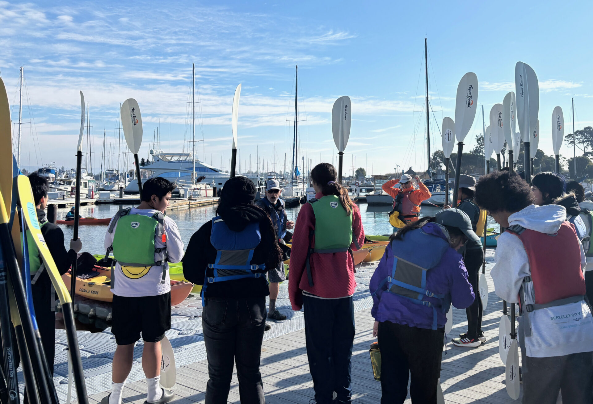 A group of high school students on a dock wearing life jackets and holding kayaking paddles