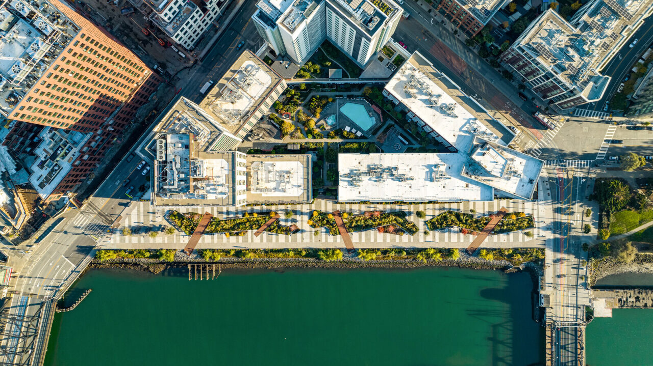 A bird's-eye view of an urban walkway/park in the center, with Mission Creek pictured below and buildings pictured above