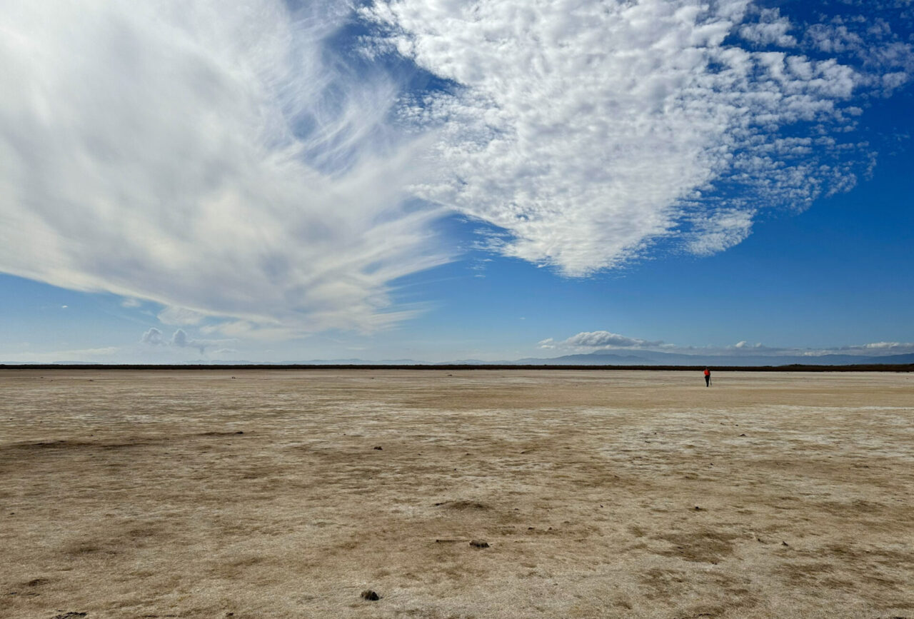 An empty, sand-colored flatland with clouds and blue sky above and one person in red walking in the distance. Mountains rise in the background