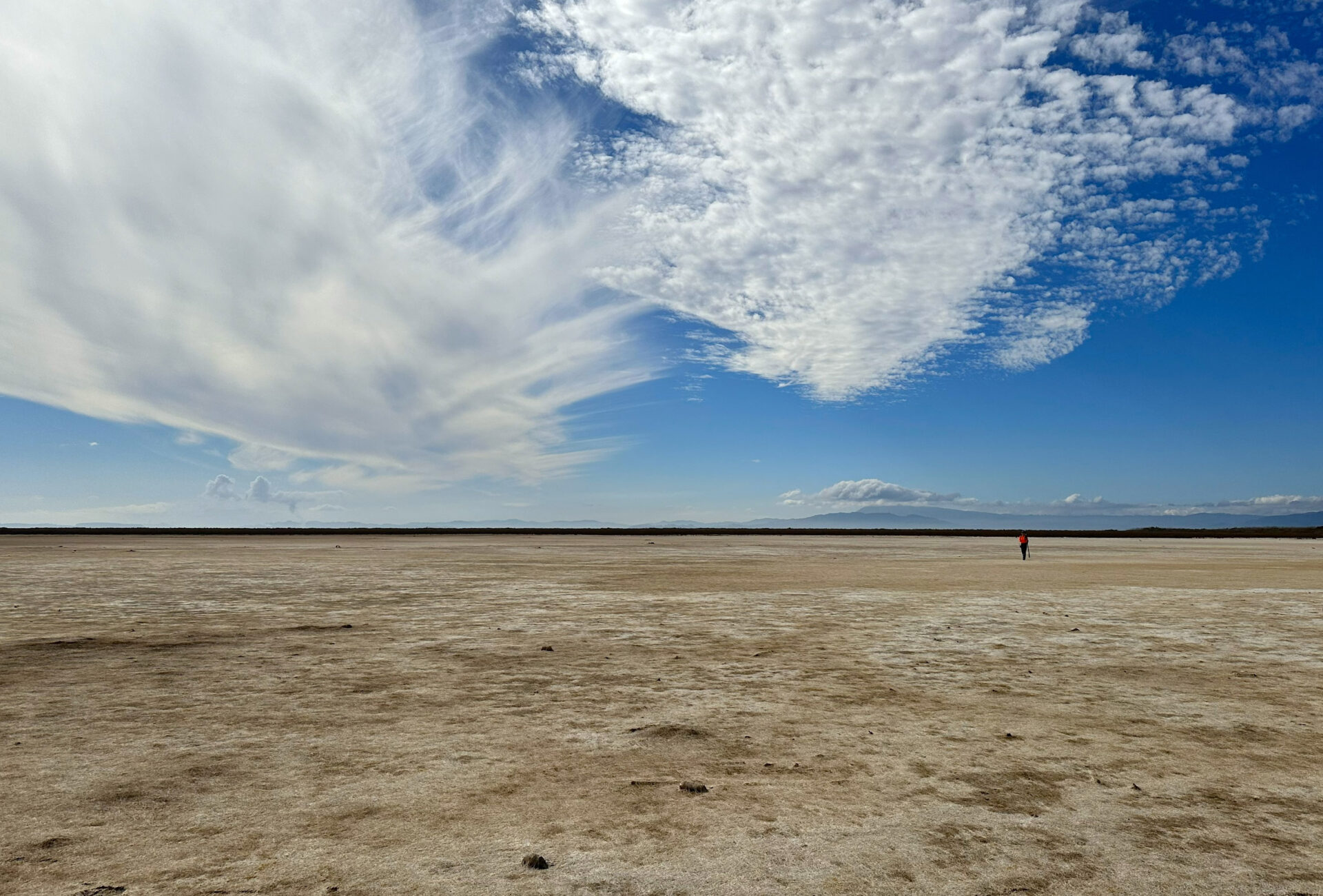 12-4-map-missionbay An empty, sand-colored flatland with clouds and blue sky above and one person in red walking in the distance. Mountains rise in the background