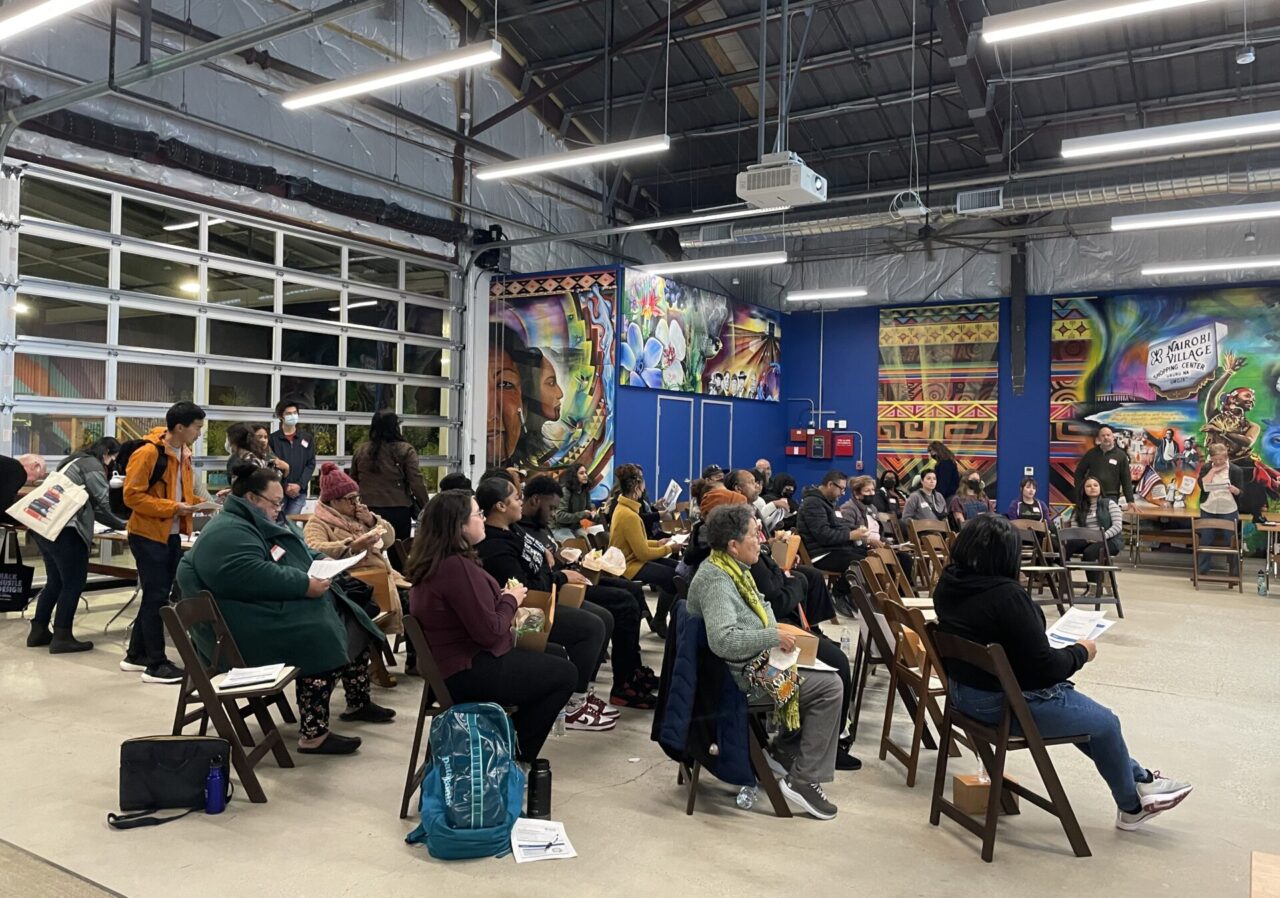 A crowd of about 30 people sits on folding chairs assembled inside a large room inside a community center