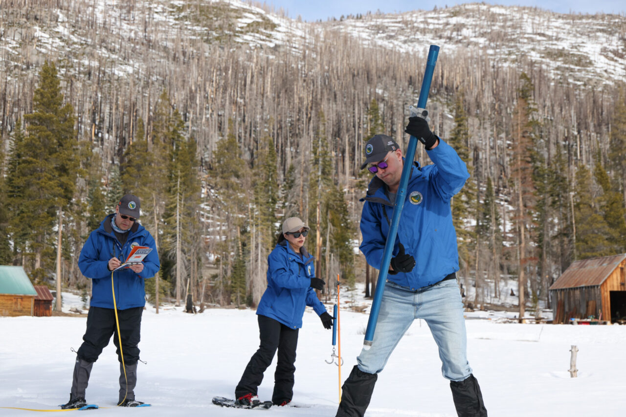 Three Department of Water Resources employees wearing blue winter jackets and snowshoes. The man on the left writes numbers in a notebook as a woman in the middle overlooks equipment and a man on the right uses a long blue cylinder to measure snow depth and water content.