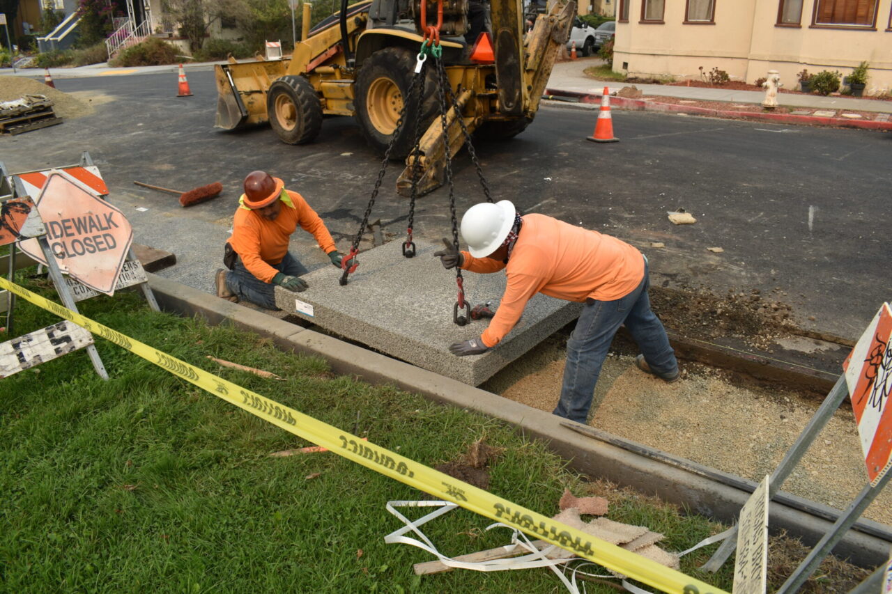 Two construction workers in orange shirts and hard hats lay down a square of pervious concrete in a parking lot with the help of a bulldozer or some other kind of construction vehicle.