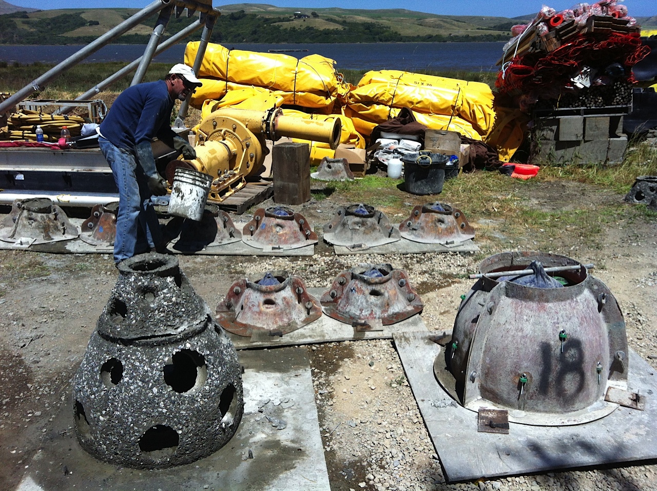 A white man wearing a white hat, sunglasses, a long sleeve blue shirt and blue jeans holds a white bucket and pours "baycrete" — a mixture of concrete, oyster shells, sand, and gravel — into molds to make reef structures for a living shoreline project. Yellow equipment is seen in the background, behind which is a body of water and land on the other side.