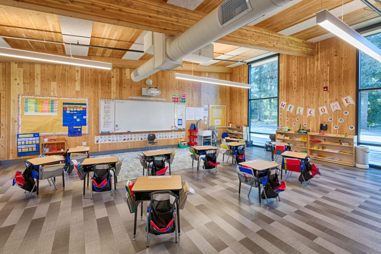 A large high school classroom with a long whiteboard in the back and bulletin boards on each side of it, with 11 individual desks arranged in front of the board. The walls and ceiling are made out of wood, and the floor is made of silver panels. There are two floor to ceiling windows on the right, with cubbies and a sign saying "welcome" in between.