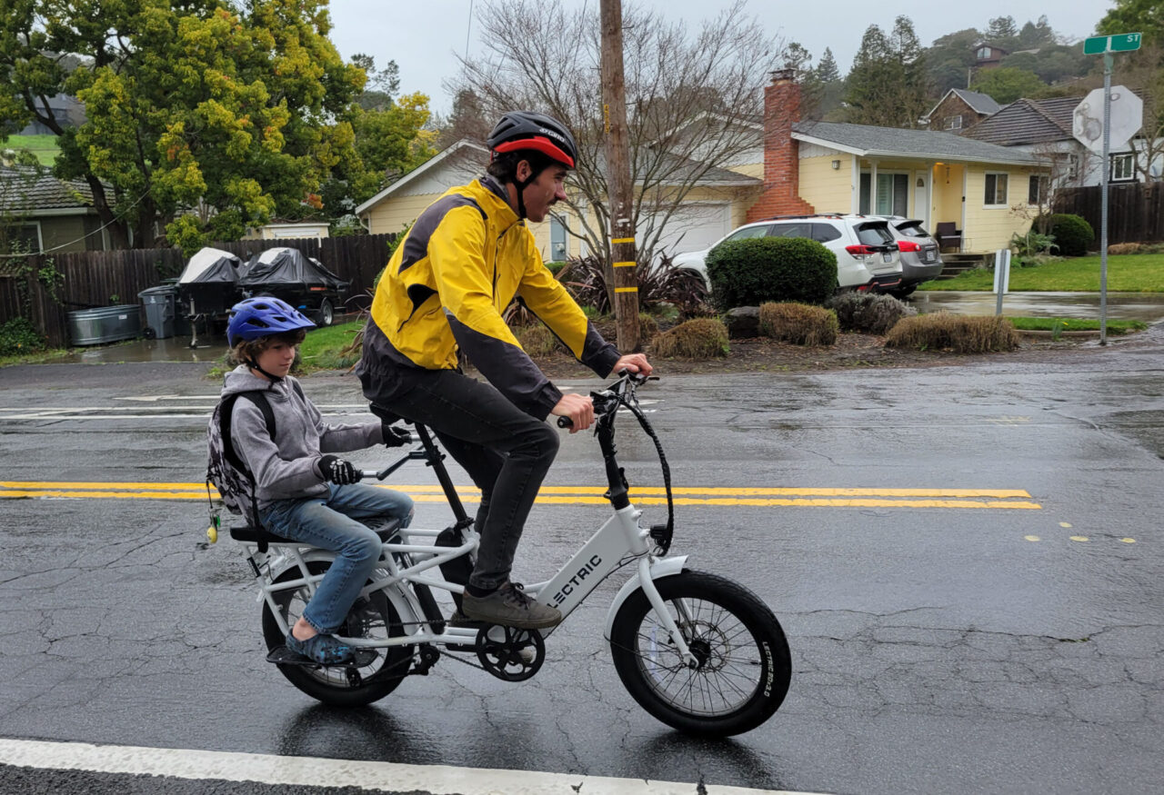 A white man wearing a yellow and black rain jacket, a black and red helmet and black pants pedals a white electric bike as his 10 year old son, wearing a blue helmet, gray zip up sweatshirt, blue jeans and a backpack, sits on the back wheel. They are riding in the right lane of a two-lane road, with a yellow house in the background and a green street sign in the top right corner of the photo
