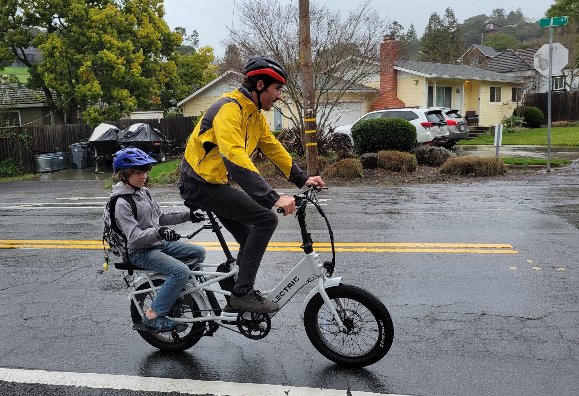 A white man wearing a yellow and black rain jacket, a black and red helmet and black pants pedals a white electric bike as his 10 year old son, wearing a blue helmet, gray zip up sweatshirt, blue jeans and a backpack, sits on the back wheel. They are riding in the right lane of a two-lane road, with a yellow house in the background and a green street sign in the top right corner of the photo