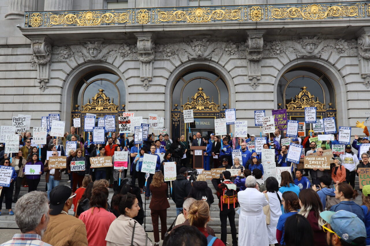 A large crowd of more than 100 people holding signs with messages like "Don't Defund Our Future," "You Can't Lay Off the Climate Crisis," and "No Climate Cuts" gather around a white man in a suit and glasses speaking at a podium in front of an ornate stone building with gold trim. Steps lead down from the demonstrators to a group of people in the foreground watching or filming the protest.
