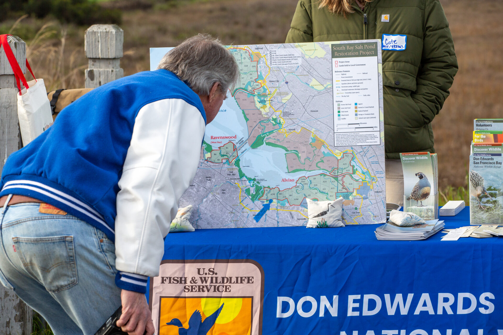 A white man with gray hair wearing a blue jacket with white sleeves and jeans leans over to look at a diagram showing salt pond restoration projects around the South Bay. The diagram is a large poster sitting upright on a U.S. Fish and Wildlife Service information table at some kind of community information session.