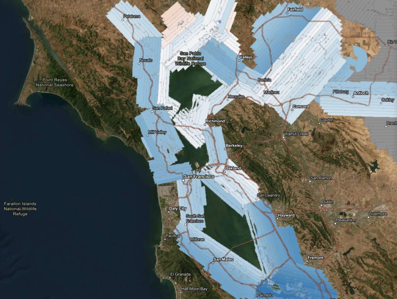 Satellite map of the San Francisco Bay Area showing overlapping LiDAR survey flight tiles displayed as semi-transparent blue and white rectangular swaths. The tiles are oriented in varying diagonal directions, reflecting different flight line azimuths used during aerial data collection. The swaths cover the bay shoreline, tidal wetlands, and surrounding lowlands extensively, spanning from Point Reyes National Seashore and Petaluma in the north, south to Half Moon Bay and Palo Alto, and east to Antioch, Concord, and Fremont. San Pablo Bay National Wildlife Refuge is labeled near the top center. A small cluster of salmon/peach-colored tiles in the upper center area contrasts with the predominantly blue swaths, possibly indicating a different data collection campaign or sensor. Open water areas of San Francisco Bay and San Pablo Bay appear dark green through the transparent overlays. Major cities labeled include San Francisco, Oakland, Berkeley, Richmond, San Rafael, Vallejo, Concord, Hayward, and San Mateo, among others. The Farallon Islands National Wildlife Refuge is noted in the lower left over the open Pacific Ocean.