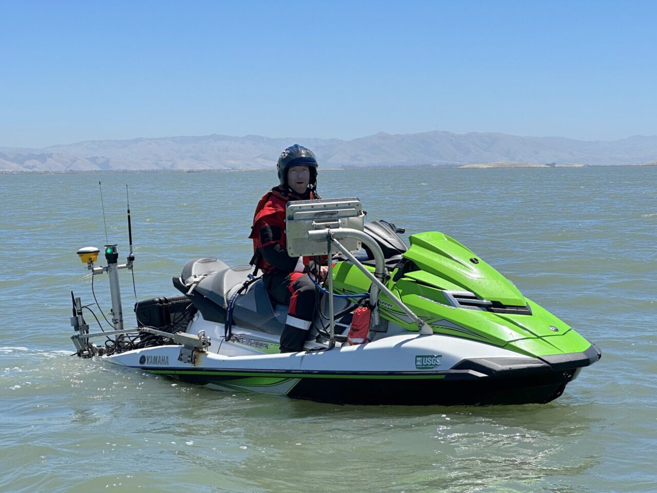 A man wearing a helmet and a red jumpsuit drives a green and white jet ski equipped with technology used to map underwater topography.