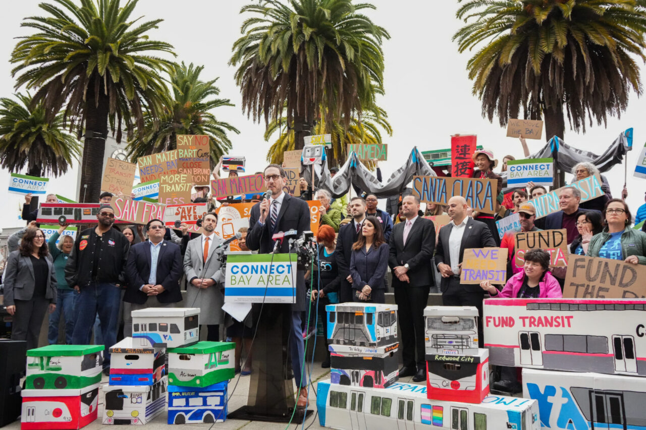 A large crowd of several dozen people holding signs and graphics in support of public transit gathered around a man in glasses and with short hair wearing a dark suit and tie speaking at a podium. Palm trees are seen overhead in the background.