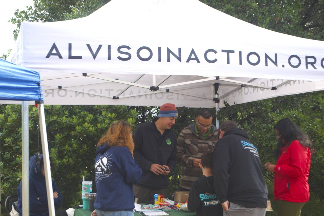 A group of seven people stand under a white tent with the words "ALVISOINACTION.ORG"
