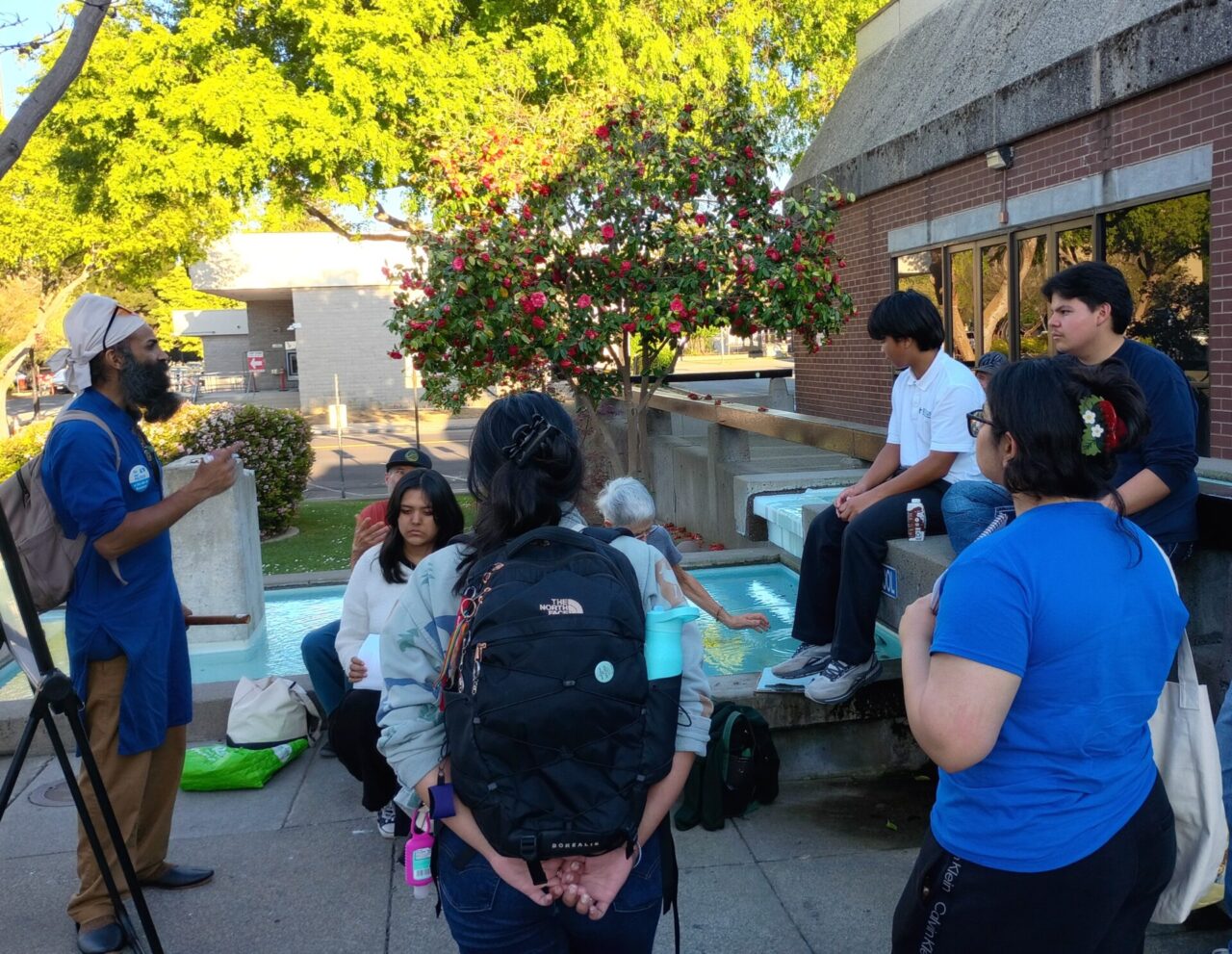 Outdoor meeting at Vallejo public library, with a man with a beard and a long blue shirt and tan pants wearing a backpack speaking to a group of eight people.